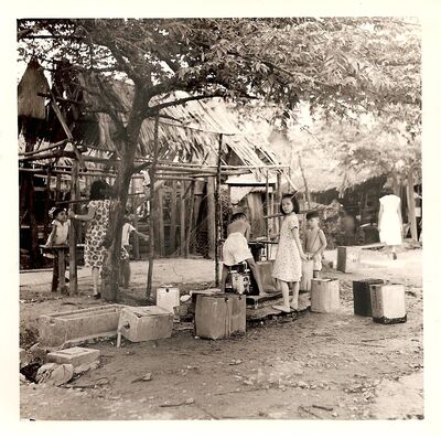 Sembawang Village - Kampung Water Point 1952
Sembawang Village located just outside the Naval Base Main Gate sprung up and prospered due to the presence of the base. The land was originally a mixture of malarial swamp and rubber plantation. In time piped water arrived but like in a lot of Singapore kampungs only by stand pipe. Even in 1958 I witnessed locals getting their water as in this photo. Collecting it in 5 gallon rectangular metal tins that were previously used for the delivery of the kerosene used for lighting & cooking.
Keywords: Kampung;kerosene;Sembawang;stand pipe